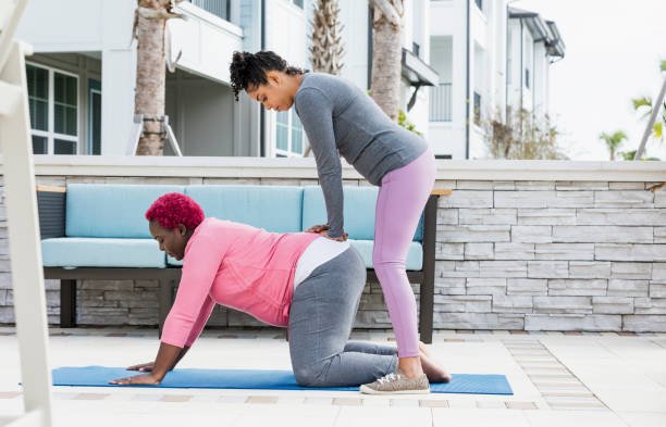 A pregnant African-American woman with her doula or birth support coach. The expectant mother, who has pink hair and is wearing a pink shirt, is on her hands and knees. The doula, a mixed race woman, is standing behind her, applying pressure to her lower back. This labor position can help ease back pain during contractions. Both women are in their 30s.